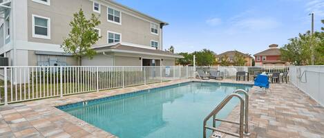 A swimming pool with a tiled deck, a white fence, and a building with multiple windows in the background.