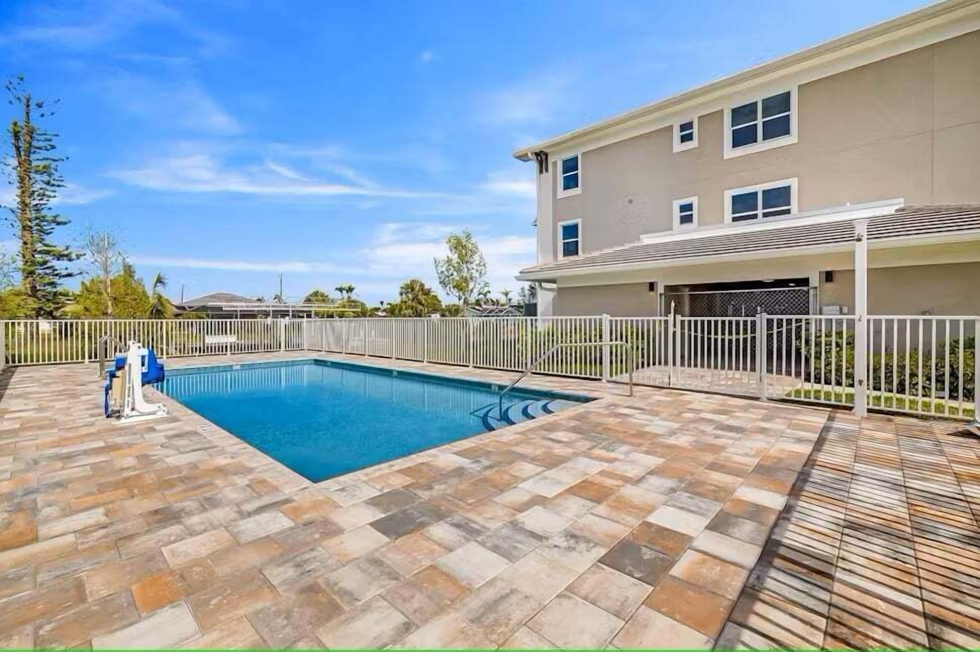 A rectangular swimming pool with a tiled deck, surrounded by a white fence and a building with multiple windows.