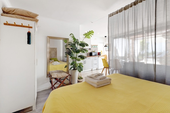 A room with a bed, a chair, a mirror, a plant, and a kitchen area.