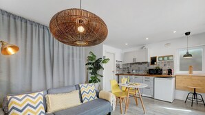 Een moderne woonkamer met een bank, een eethoek met gele stoelen, een keuken met witte kasten, en een hangende rotan lamp.