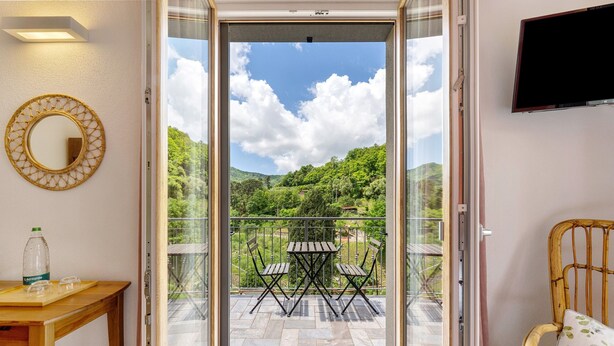 Une porte coulissante s’ouvrant sur un balcon équipé d’une table et de chaises, d’un fauteuil en osier et d’un téléviseur fixé au mur.