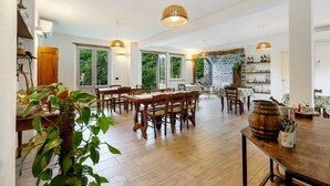 Une salle à manger spacieuse, avec des tables et des chaises en bois, de grandes fenêtres et un mur en pierre.