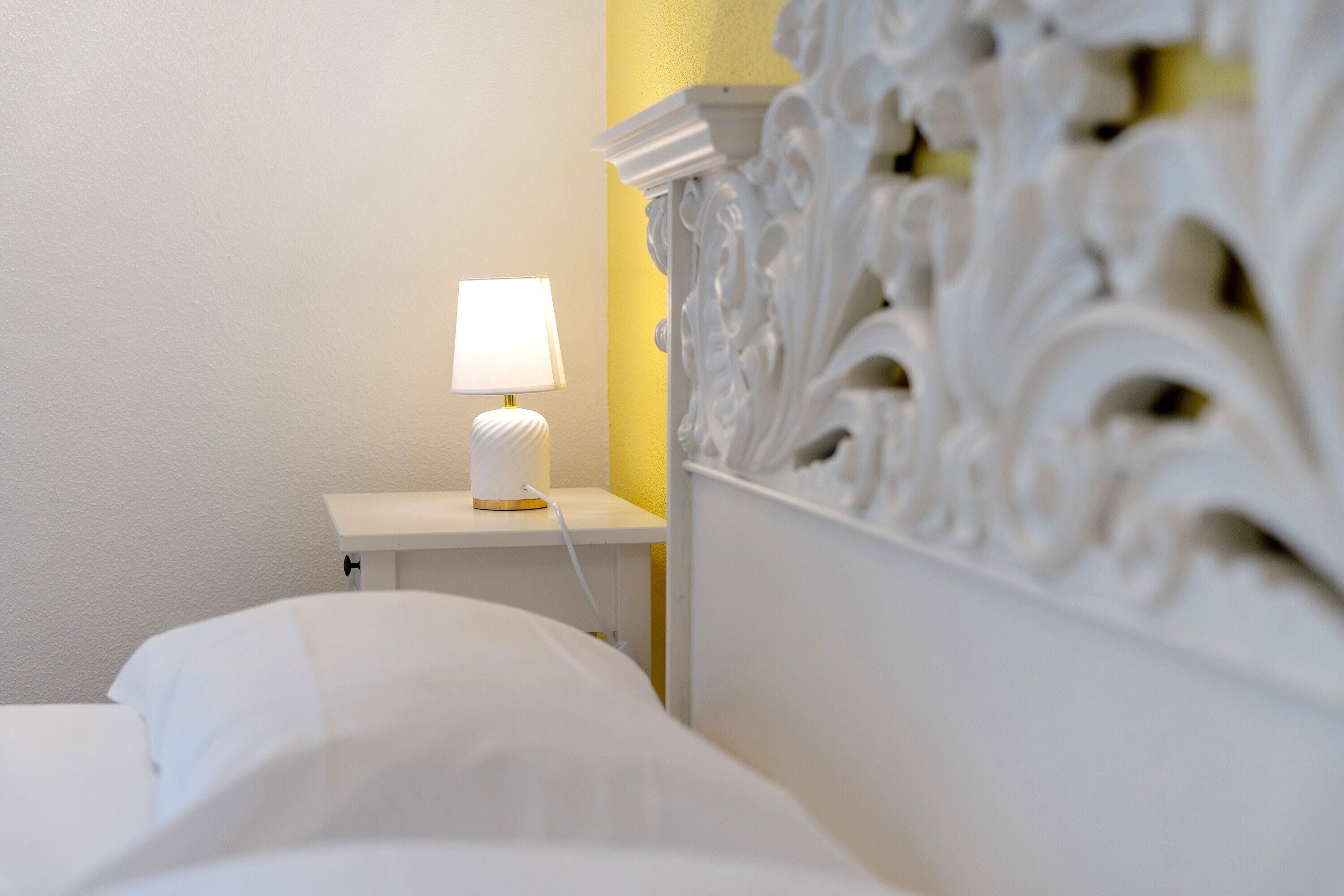 A bed with white bedding, a bedside lamp, and a white nightstand with a yellow wall in the background.