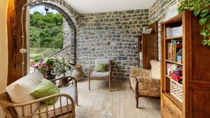 A cozy room with stone walls, a wooden bookshelf, wicker furniture, and a view of greenery outside.