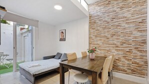 A modern living room with a sofa, a dining table with chairs, a small garden visible through a sliding door, and a brick-patterned wall. - Apartment 'Lisboa Sweet For Familys' with Private Terrace, Wi-Fi and Air Conditioning (Lisboa)