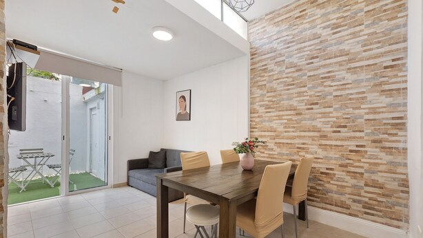 A dining area with a wooden table, four chairs, a vase with flowers, a grey sofa, and a wall with a brick pattern. - Apartment 'Lisboa Sweet For Familys' with Private Terrace, Wi-Fi and Air Conditioning (Lisboa)
