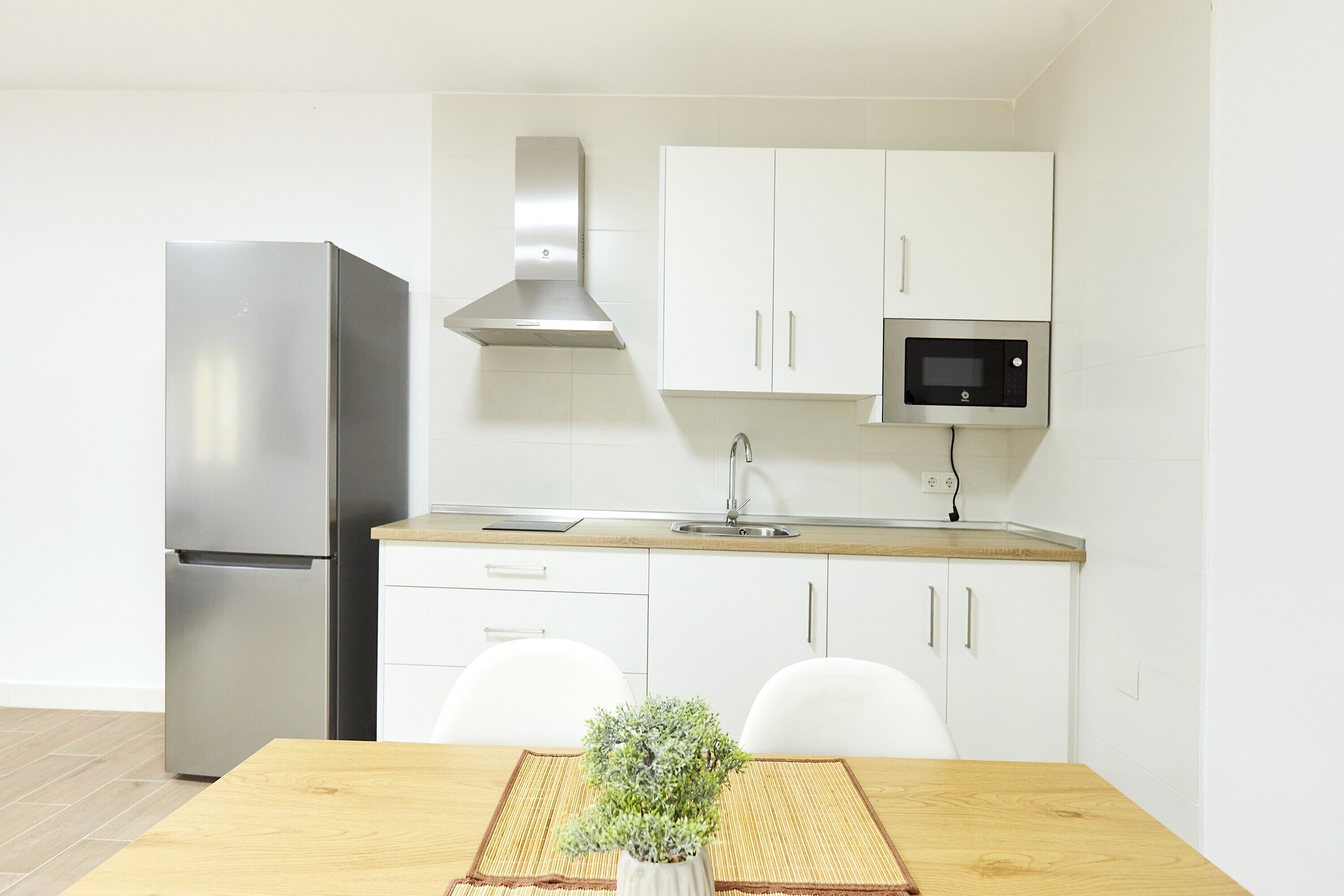 Una cocina moderna con armarios blancos, un refrigerador de acero inoxidable, un microondas y una mesa de comedor de madera con una planta en maceta.