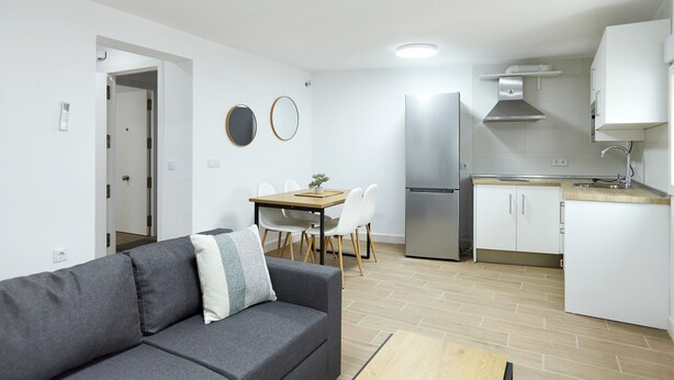 Un appartement moderne avec un canapé gris, une table à manger avec des chaises, un coin cuisine équipé d’un réfrigérateur et de placards, et un couloir menant à une autre pièce.