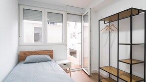 Une chambre à coucher comprenant un lit, une petite table de chevet, une armoire avec des étagères et une porte-fenêtre coulissante donnant sur un balcon.