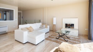 Een moderne woonkamer met een witte bank, een glazen salontafel, een flatscreen televisie en een groot raam.