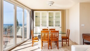 Een eetgedeelte met een glazen tafel en stoelen, een wit kastje met glazen deuren, en uitzicht op de zee door grote ramen.