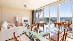Een moderne woonkamer met een glazen eettafel, houten stoelen, een witte bank en een groot raam met uitzicht op zee.