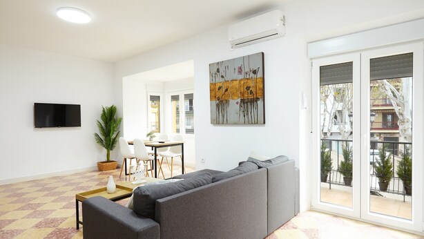 Ein Wohnzimmer mit einer Couch, einem Fernseher, einem Essbereich und einem Balkon mit Blumentöpfen.