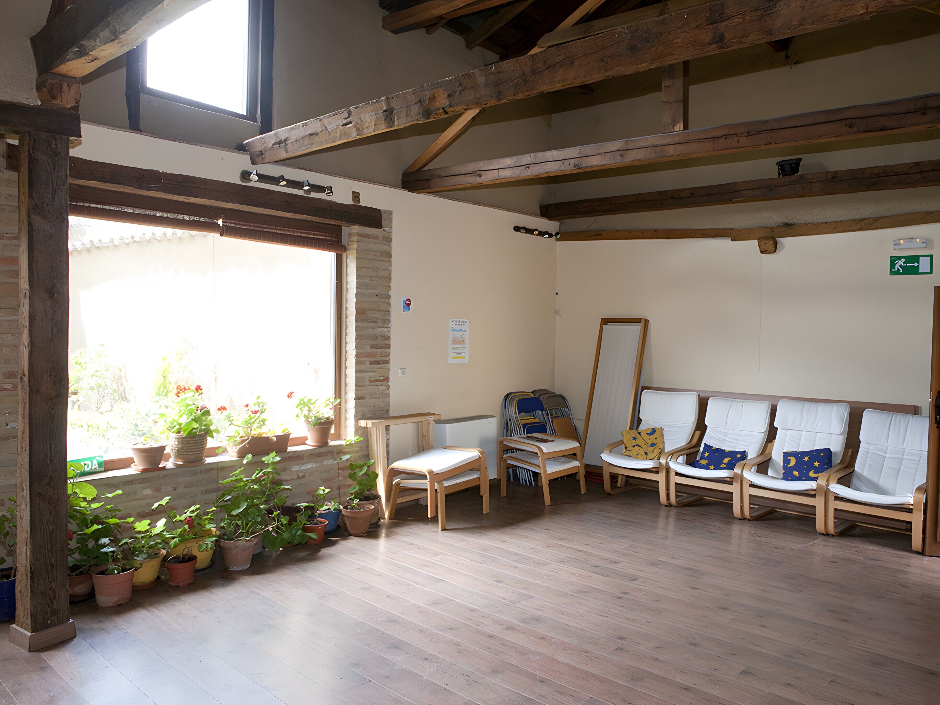 Una habitación con vigas de madera, una ventana con plantas en macetas y una fila de sillas.