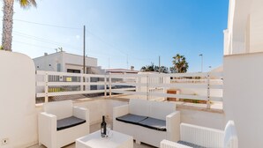 Una terraza en la azotea con muebles de mimbre blanco, una mesa con una botella y un vaso, y vistas a edificios y palmeras.