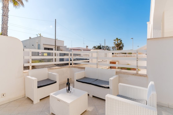 A rooftop patio with white wicker furniture, a table with a bottle and glass, and a view of buildings and palm trees.