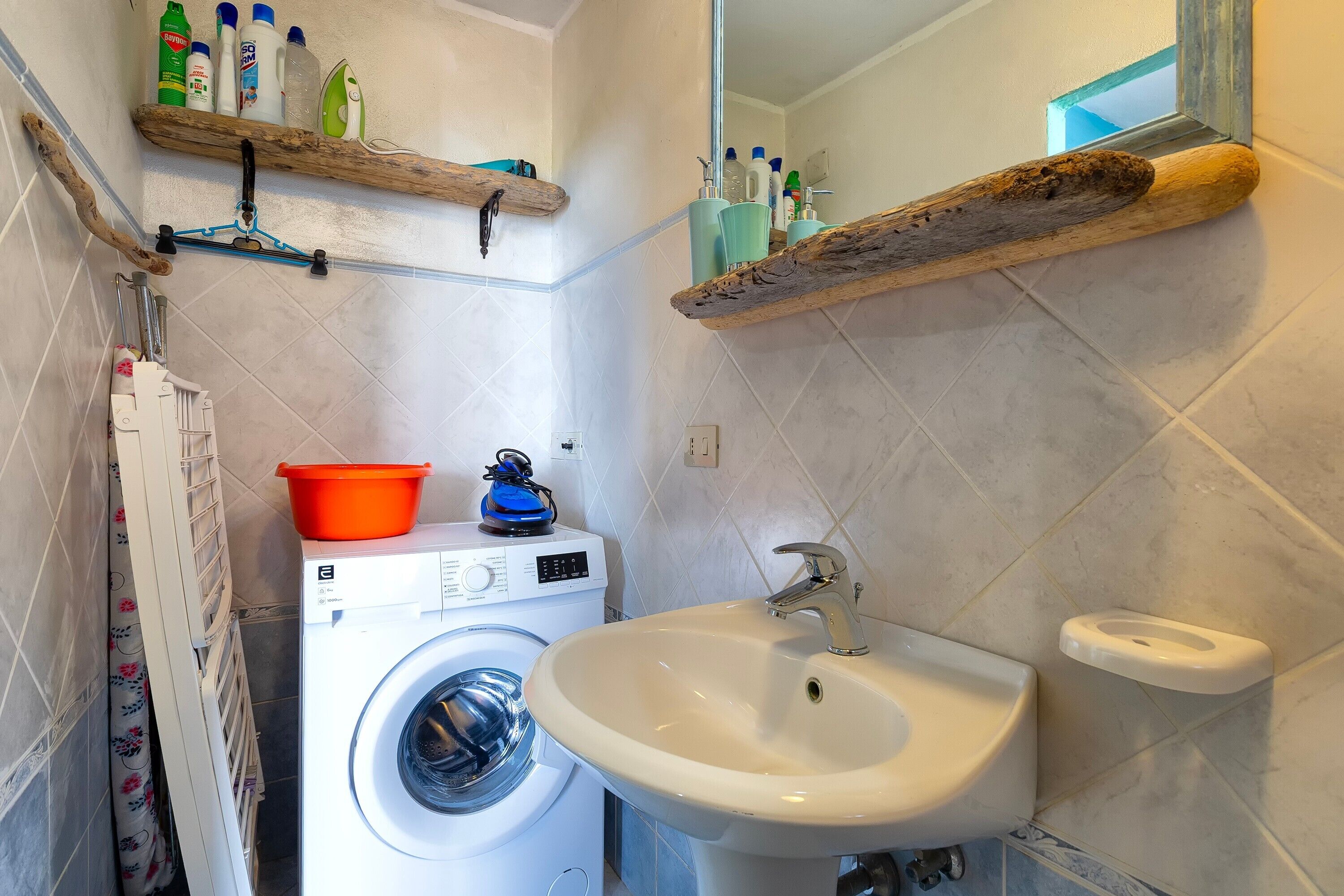 A compact bathroom with a washing machine, a sink, and wooden shelves holding toiletries.