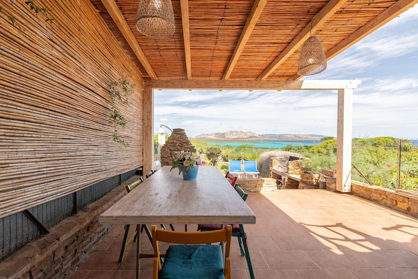 Un área de comedor al aire libre con una mesa y sillas de madera, un muro de piedra y vistas al mar y a las montañas.