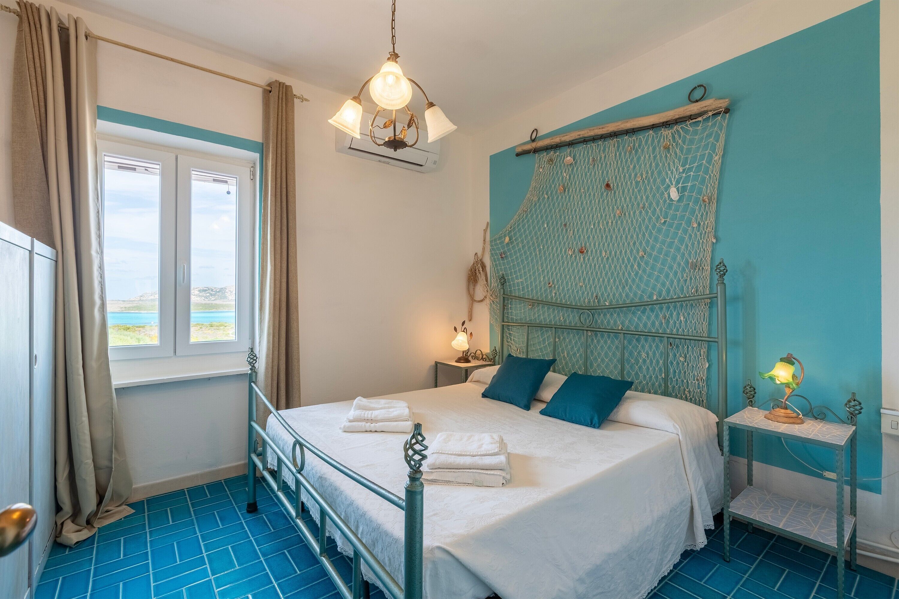 A bedroom with a bed, a nightstand, a lamp, a window with curtains, and a view of the beach.