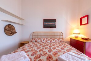 Een netjes opgemaakt bed met een bedsprei met patroon, een aan de muur hangend schilderij, een bedlampje en een klein houten nachtkastje.