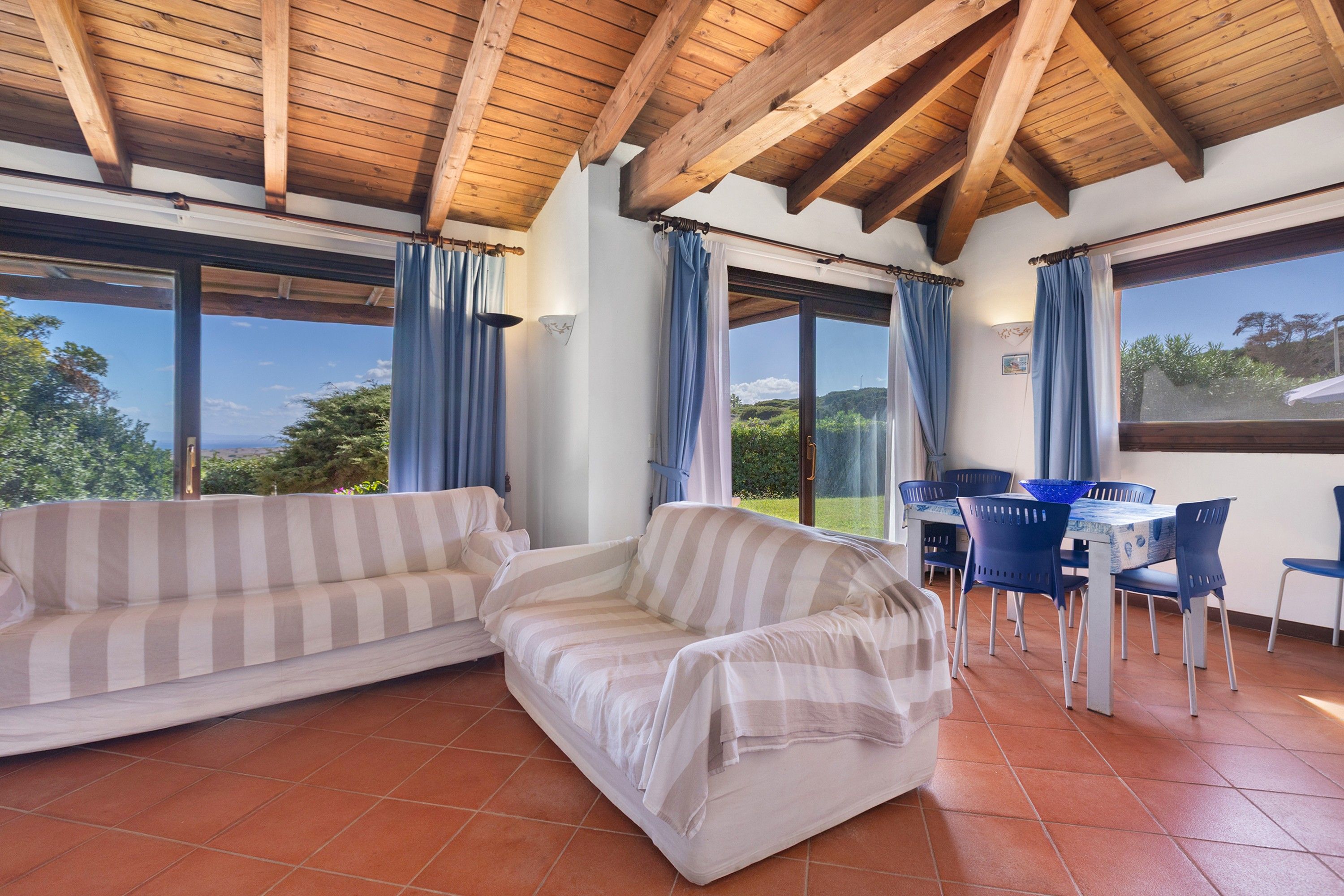 A room with a wooden ceiling, a striped sofa, a dining table with chairs, and large windows with curtains.