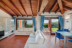 A spacious living area with a wooden ceiling, a white sofa, a TV, and a dining table with blue chairs.
