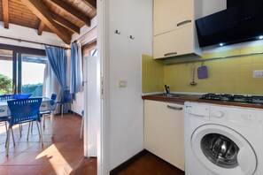 A compact kitchen with a washing machine, a dining area with blue chairs, and a view through a glass door.