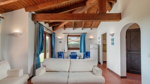 A living room with a sofa, dining table with chairs, wooden ceiling beams, and a view through a window. - Villa 'Alba Piscina Vista Mare' with Sea View, Wi-Fi and Air Conditioning (Stintino)