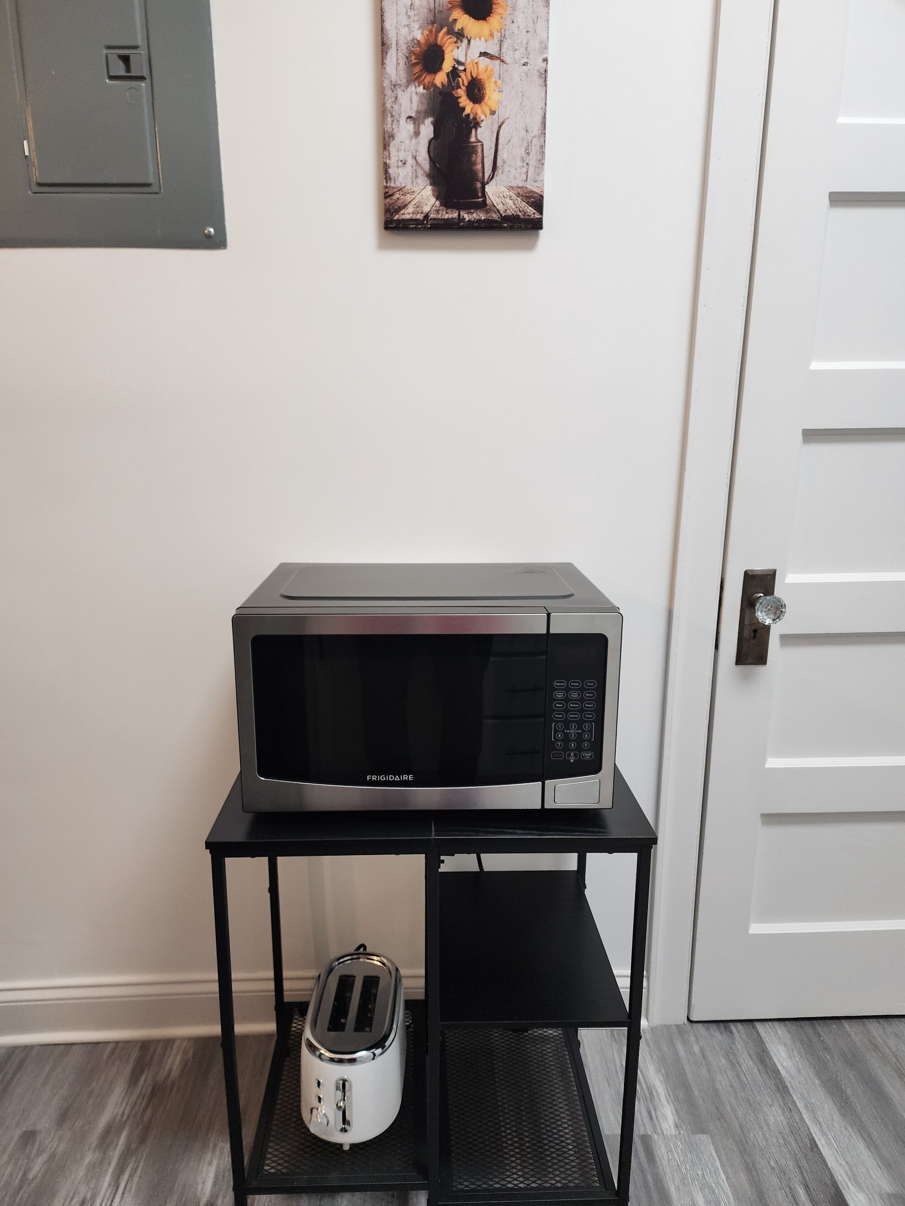 A kitchen area with a microwave on a black metal stand, a toaster, and a painting of sunflowers on the wall.