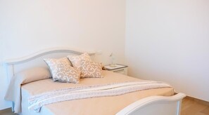 Uma cama com cabeceira branca, duas almofadas estampadas, uma mesa de cabeceira com abajur e um cobertor.