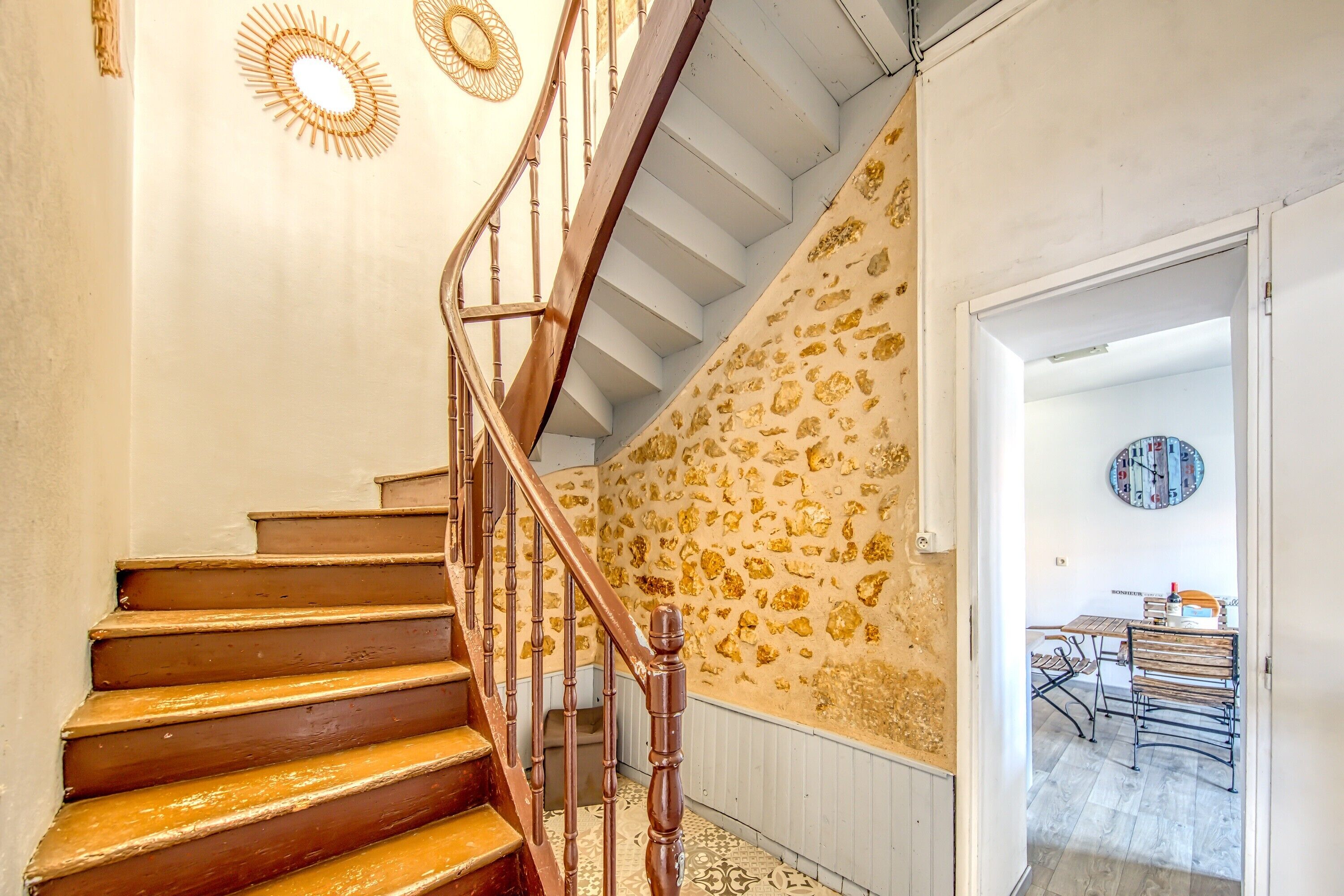 A staircase with wooden steps and a metal railing, leading to a room with a patterned wall and a dining area with a table and chairs.