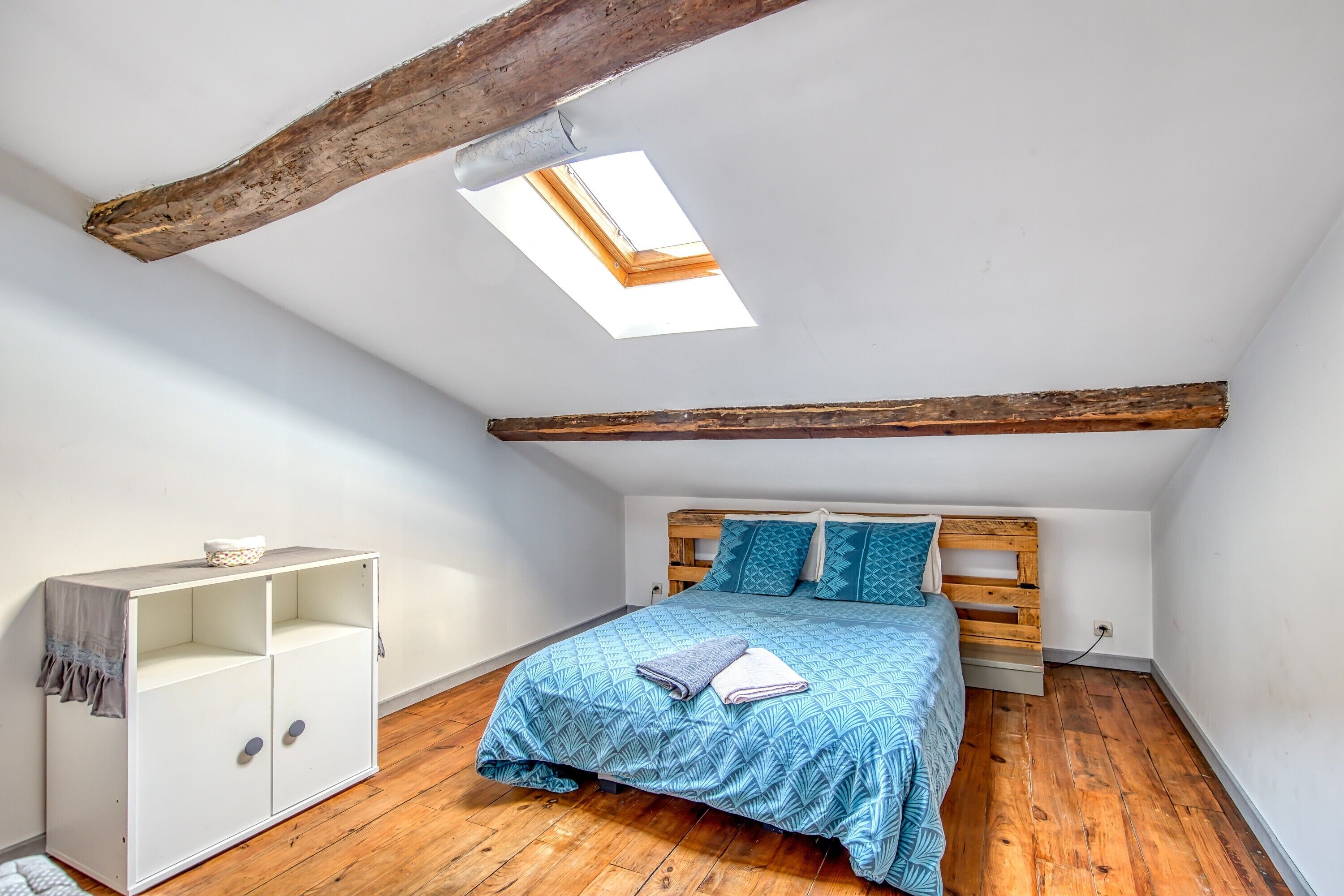 Un dormitorio con techo inclinado, tragaluz, cama con colcha azul estampada, cabecera de madera y aparador blanco con cajones.