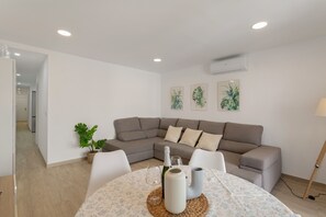 A modern living room with a grey sectional sofa, a round dining table with a woven placemat, and a potted plant. - Vacation Apartment 'Lupama Suite' with Private Pool, Wi-Fi, and Air Conditioning (Argueniguín)