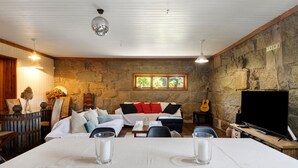 A cozy living room with a stone wall, a sofa set, a dining table, and a television. - Holiday Home 'Casa Chocolate' with Mountain Views, Private Pool and Wi-Fi (Porto)