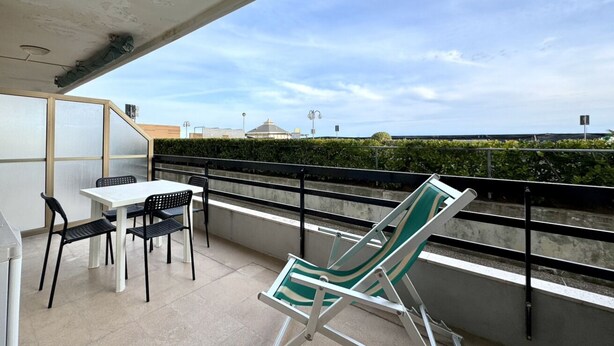 A balcony with a table and chairs, a green and white striped lounge chair, and a view of the sea and distant buildings.
