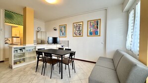 A modern living room with a grey sofa, a dining area with a table and chairs, a kitchen with a refrigerator and a TV, and artwork on the walls.