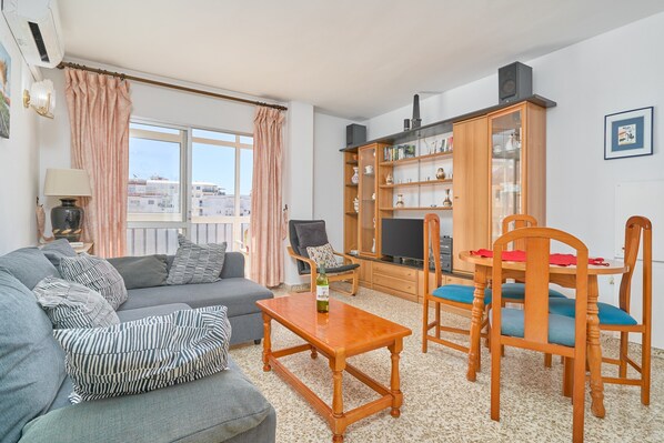 A living room with a sofa, a coffee table, a TV stand, and a dining area with chairs.