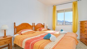 Une chambre avec deux lits, une tête de lit en bois, une fenêtre avec des rideaux jaunes, une commode en bois et une table de chevet avec une lampe.
