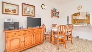 Un coin repas avec une table et des chaises, un téléviseur posé sur un meuble en bois, des tableaux encadrés au mur et un coin cuisine avec une horloge.
