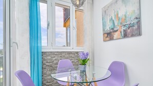 Uma sala de jantar com mesa de vidro, cadeiras roxas, um vaso com flores e um quadro na parede.