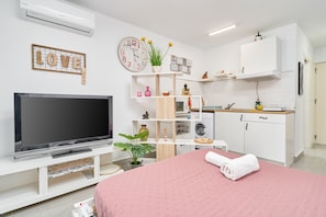 Una sala moderna con televisor de pantalla plana, una cama rosa, un sofá blanco y una cocina con armarios blancos, además de un reloj montado en la pared.
