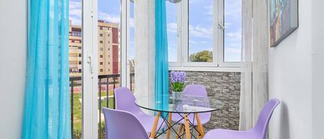 A room with a glass table, purple chairs, a hanging light fixture, and a view of buildings through large windows.