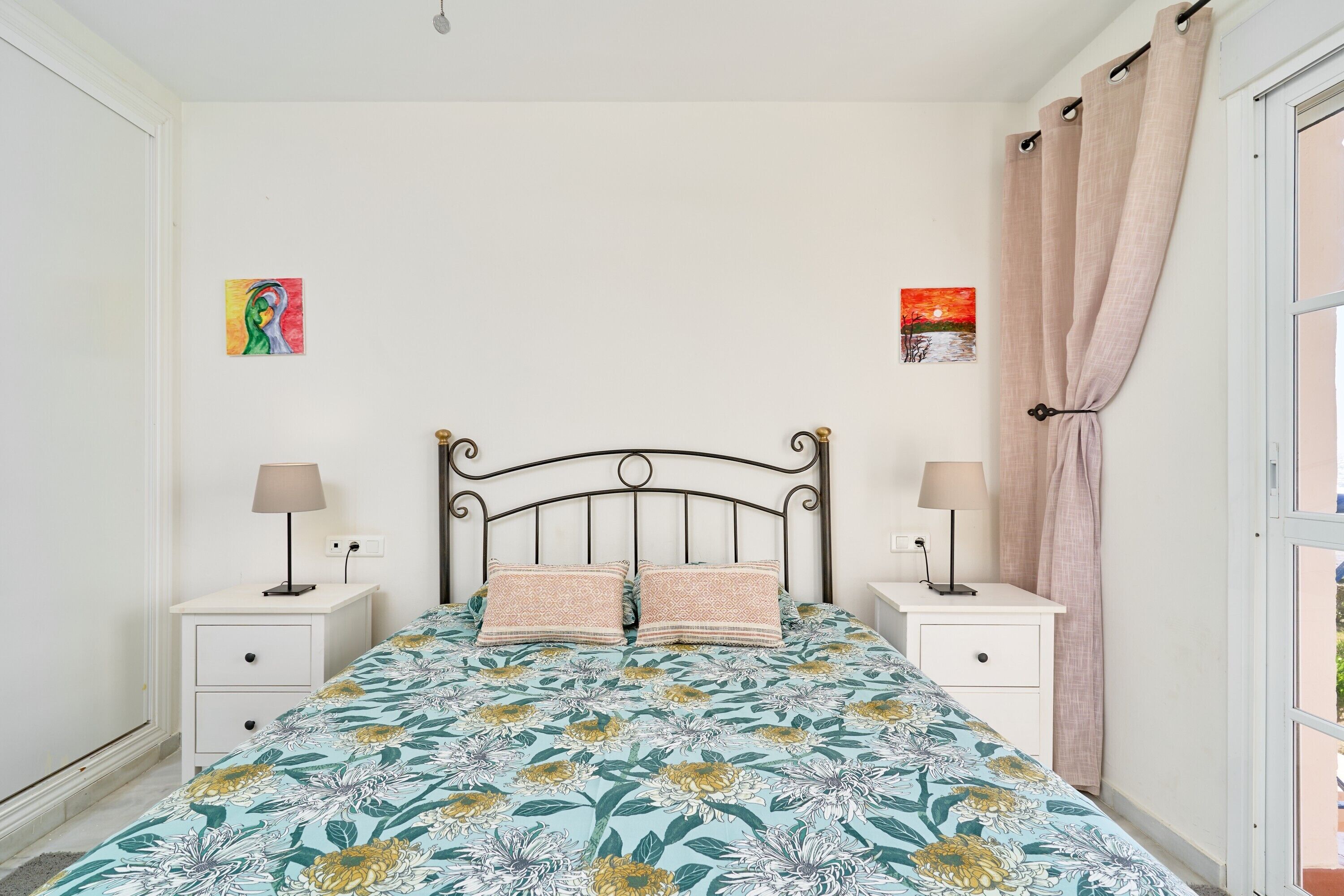 A bedroom with a bed, two bedside tables, a nightstand, a window with curtains, and two wall-mounted paintings.