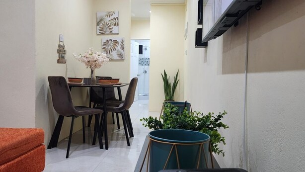 Un pasillo estrecho con un comedor que incluye una mesa, sillas y un jarrón de flores. Hay una planta en maceta y un tambor en primer plano.