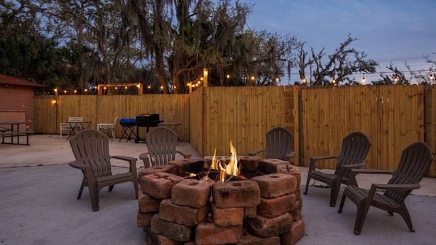 A backyard with a brick fire pit surrounded by Adirondack chairs, a wooden fence, and string lights.