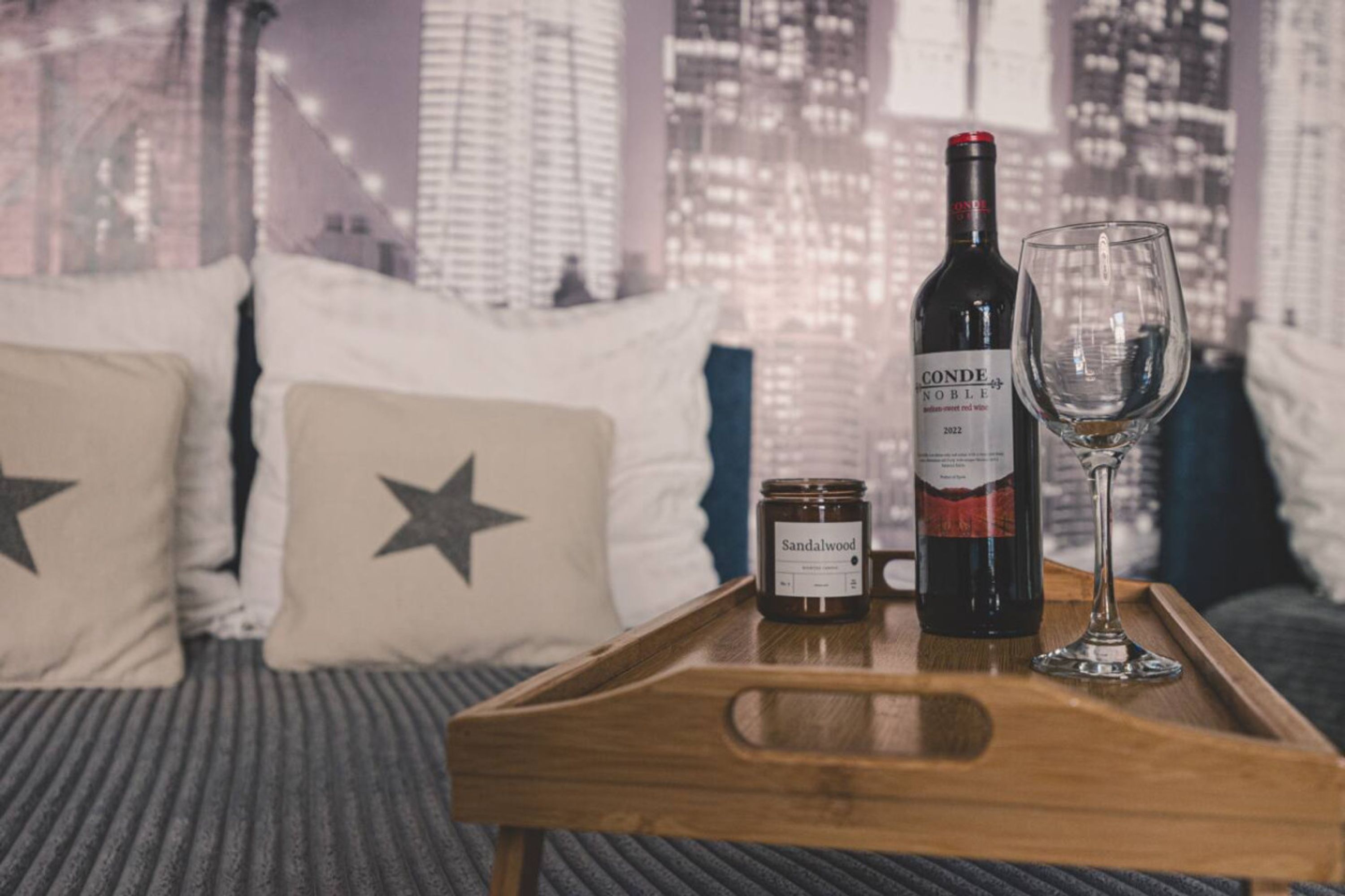 Una bandeja de madera con una botella de vino Conde Noble 2022, una copa y una vela sobre una cama con almohadas estampadas con motivos de estrellas.