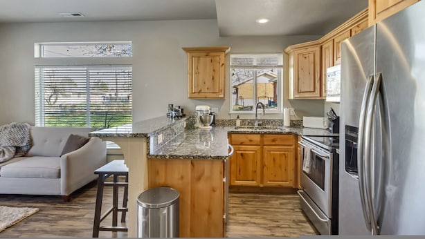 A modern kitchen with wooden cabinets, a stainless steel refrigerator, granite countertops, and a built-in island with bar stools. - Hill Field 2 by CDH in Layton (Layton)