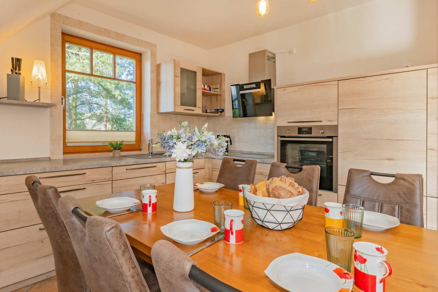 Une cuisine moderne avec des meubles en bois, une table à manger pour quatre personnes et une fenêtre donnant sur un paysage arboré.