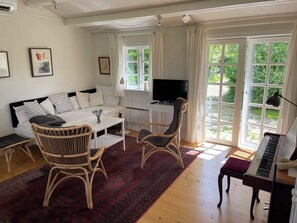 Ein Wohnzimmer mit einer Couch, zwei Korbstühlen, einem Couchtisch, einem Fernseher, einem Klavier und einem großen Fenster mit Vorhängen.