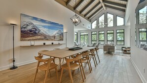 A spacious dining area with a long wooden table and chairs, large windows, and a mountain landscape painting on the wall.